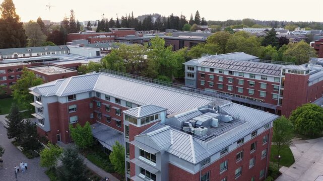Red brick university campus buildings with metal rooftops, tree-lined walkways, students on paths, and forested hillside under soft evening light. Eugene, Oregon, USA. Aerial flyover shot. Quiet scene