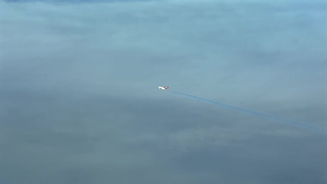 right side aerial cockpit view of a twin-engine massive jet airplane with a red tail, leaving a fading contrail over a layer of ethereal clouds