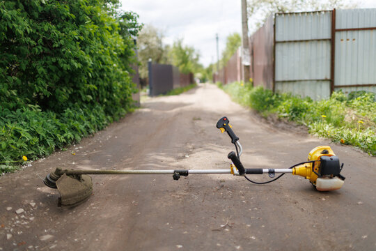 A ground-level shot of a yellow string trimmer lying horizontally across a dirt village lane with selective focus