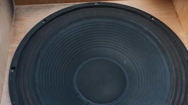 A close-up view of a large black subwoofer speaker cone vibrating intensely. The paper acoustic diffuser operates at low bass frequencies inside a raw wooden cabinet enclosure.