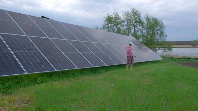Farmer checking solar panels performance while working in the garden A man with a hoe stands near home solar panels, calculating energy efficiency on a smartphone and taking notes in a notebook. 