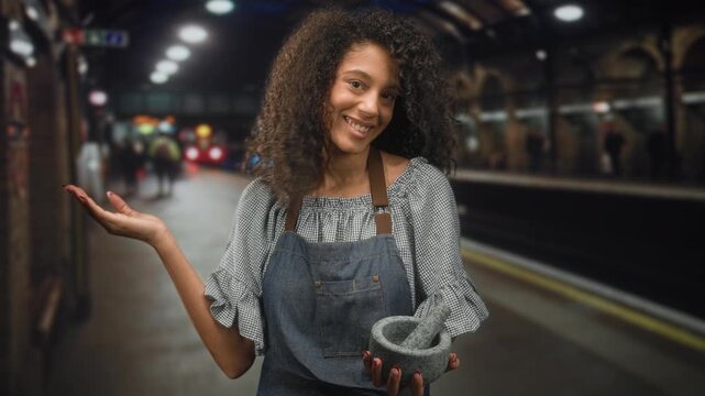 Woman holding mortar and pestle with palm up gesture on subway platform inside building; joy community.