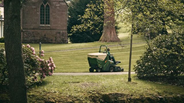 Groundskeeper driving a large green ride-on collector mower on a paved path near a historic stone chapel with a gothic window in a lush green park.