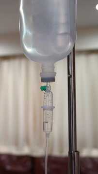 A close-up shot of an intravenous (IV) drip infusion set with saline solution drops in a hospital room, featuring a blurred background.