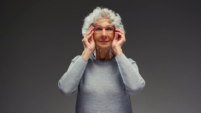 Elderly woman puts on glasses on her head. Old female wear plastic eyeglasses, taking care of eyesight. Close-up portrait, senior lady adjusting or putting spectacles, calm everyday scene. Studio