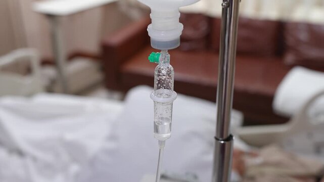 A close-up shot of an intravenous (IV) drip infusion set with saline solution drops in a hospital room, featuring a blurred background.