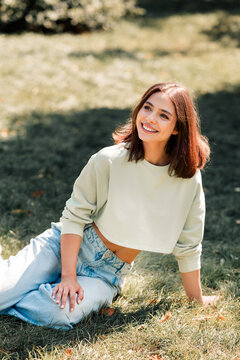 Young woman smiling and relaxed sitting on grass in park wearing cropped sweatshirt and jeans enjoying sunny outdoor moment