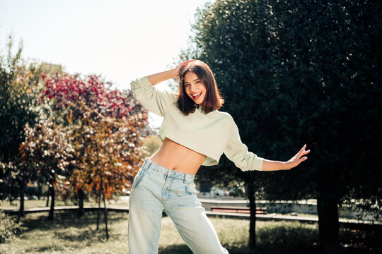 Smiling young woman in cropped sweater and jeans poses playfully outdoors in a sunny park