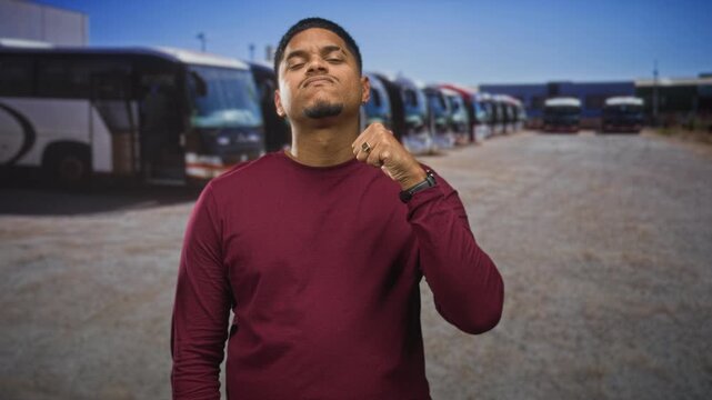 Man with beard in maroon sweater giving thumbs up gesture on a street lined with parked coaches and buses; confidence.