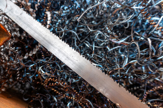 Serrated hacksaw blade lies across tangled curled metal shavings with blue and rusty tones, showing cutting, workshop waste, machining detail, and industrial metalworking texture.