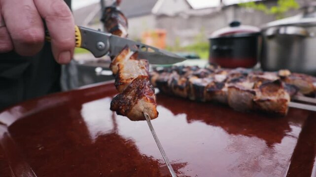 A person uses a folding pocket knife to slide a piece of hot, grilled meat from a metal skewer onto a large brown tray. The meat features visible char marks from an outdoor barbecue grill.