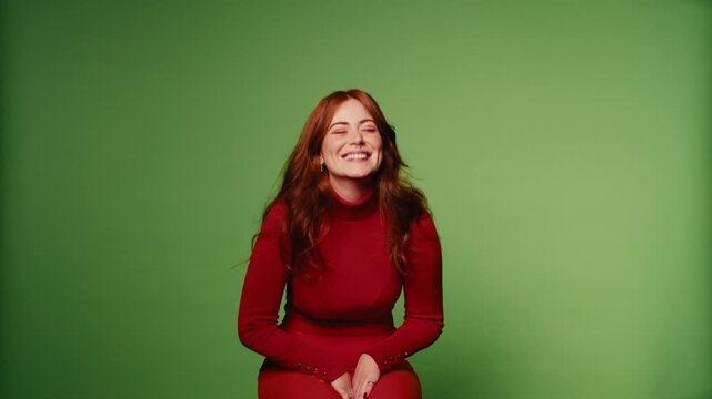 A happy young woman with long red hair sits against a green screen, laughing and smiling.