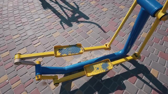 Outdoor exercise equipment is installed on a multicolored stone pavement. The metal elliptical trainer features yellow arms and blue support poles. Shadows fall across the sunny ground.