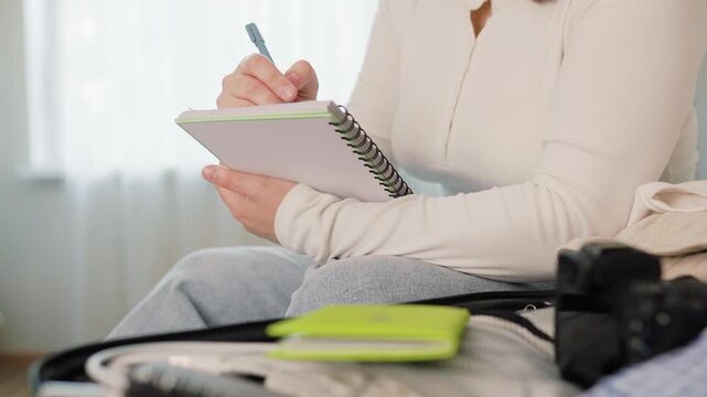 Woman doing travel preparation for a summer vacation. She writes a packing list in a notebook and packs clothes into her luggage, enjoying the vacation mood. High quality 4k footage