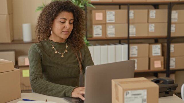 Woman rubbing neck while typing on laptop amid parcel boxes at desk in building; small business stress.