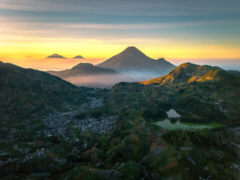 Aerial view of the Dieng plateau featuring terraced fields, a mountain village, and volcanic peaks under a golden sunrise sky in Kejajar, Central Java, Indonesia.