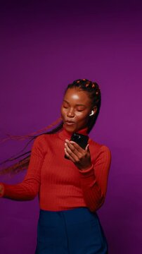 Energetic black woman with long braids enjoys music on her phone and dances against a vibrant purple backdrop in a vertical medium shot.