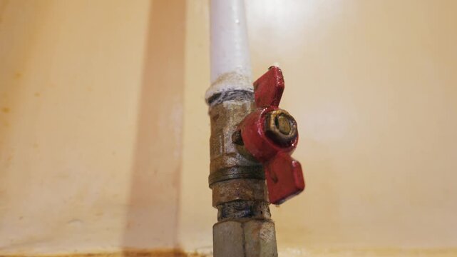 Red ball valve sits on a weathered pipe showing signs of rust and heavy wear against a beige wall in a tight macro shot. Concept of maintenance, aging infrastructure, and industrial decay.