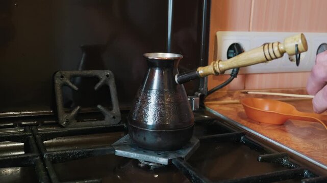 A traditional copper cezve rests on a lit gas stove burner for brewing coffee. A hand holds the wooden handle to control the heat. The cozy kitchen background features orange details.