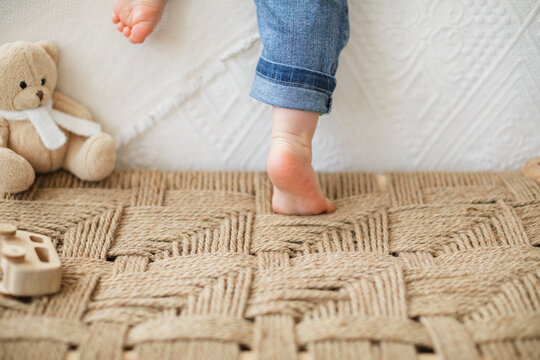 Close-up rear view of a barefoot male toddler in jeans standing on a woven bench climbing onto a bed