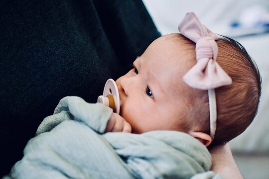 Close-up of a woman holding a newborn baby girl wearing a bow in her hair and sucking on a pacifier wrapped in a blanket