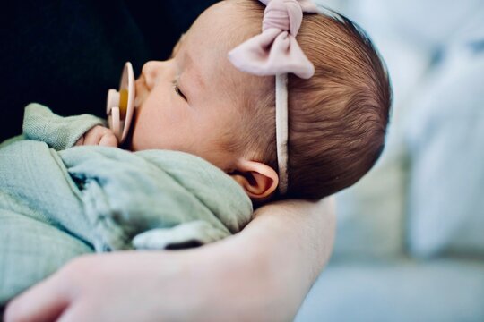 Close-up of a woman holding a sleeping newborn baby girl wearing a bow in her hair and sucking on a pacifier wrapped in a blanket