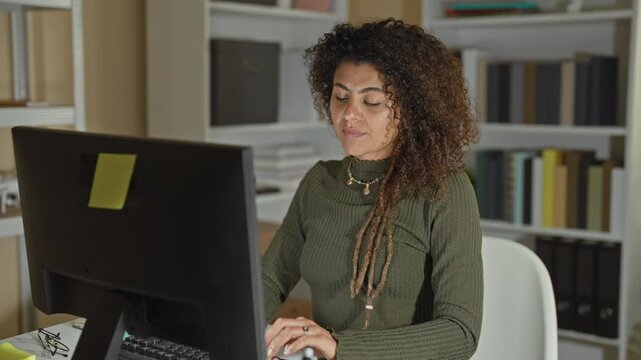 Woman rubs shoulder while using computer and mouse at desk in office building with monitor and keyboard visible; fatigue neck strain.