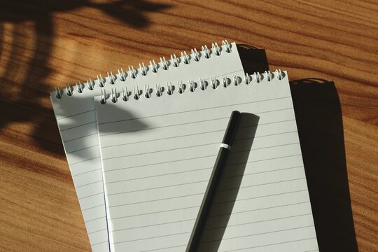 Close-up overhead view of a pencil on two Blank lined spiral note pads on a wooden desk in sunlight