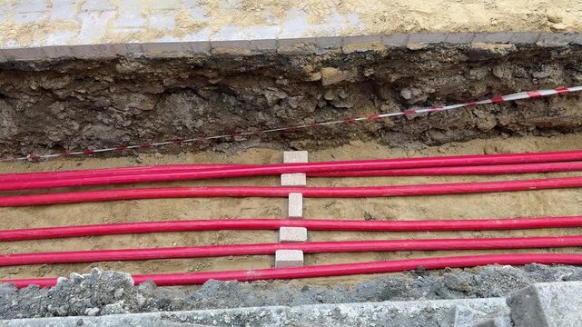 Protective red pipes lined on a sand bed inside an open soil trench at an urban site. Detail of electricity, network and telecom infrastructure work.