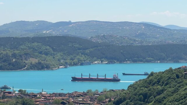 Red and black hulled bulk carrier ship sailing safely in the Bosphorus. Panoramic landscape shot of forested hills, residential areas, and maritime transport.
