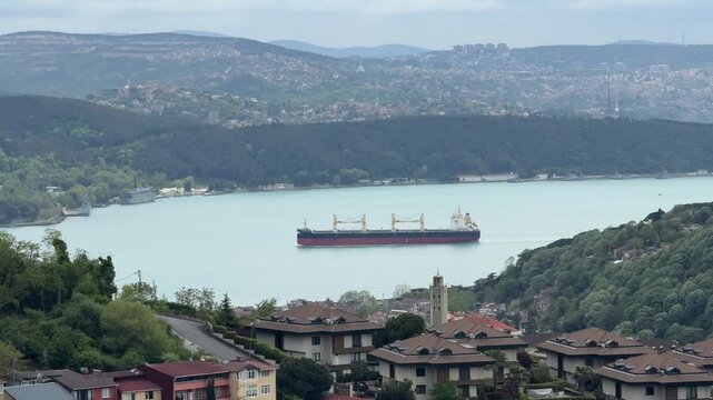 Red and black hulled bulk carrier ship sailing safely in the Bosphorus. Panoramic landscape shot of forested hills, residential areas, and maritime transport.