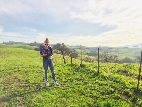 Young woman in ripped jeans standing in the countryside looking at her mobile phone, Pleasanton, California, USA