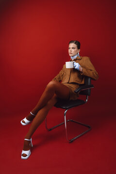 Close-up of a serious woman sitting in a chair drinking a mug of coffee in front of a red background