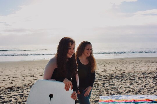 Two smiling young woman standing on a beach with a surfboard, Rio Del Mar, California, USA
