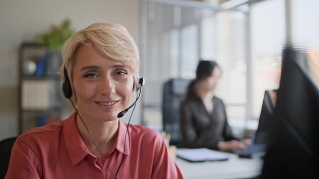 Chest up portrait of cheerful female adult operator in headset smiling at camera while working in modern call center office