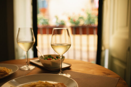 Close-up of two glasses of white wine on a dining table with two plates of pasta and salad by an open patio door in summer
