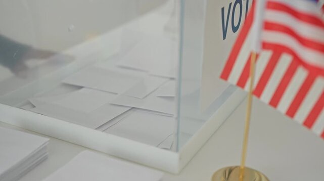 Woman inserts a folded ballot into a clear ballot box beside small us flags on a polling table in a building; patriotism civic duty.
