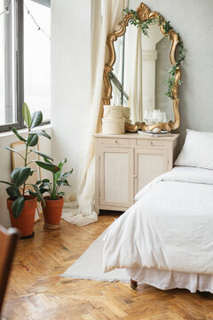 Vintage style bedroom with a gold framed mirror on a wooden dresser next to a double bed and figus houseplants