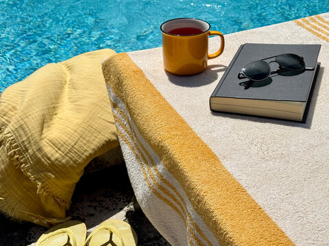 Close-up of a cup of tea, book and sunglasses on a sun lounger by a swimming pool