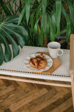 Plate of Freshly baked Croissants filled with cherries and a cup of coffee on a woven bench by monstera and palm houseplant