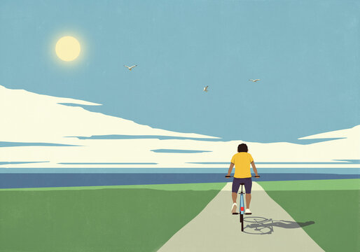 Young man riding bicycle on bike path toward sea under sunny blue sky