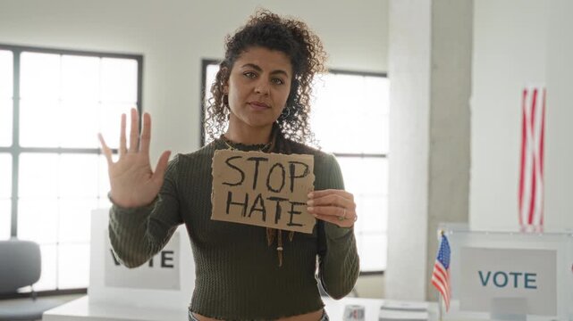 Woman holding cardboard sign reading stop hate while showing palm out toward camera inside a voting building with vote booths and us flags visible; solidarity protest.