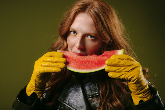 Close-up portrait of a redhead woman wearing gloves and eating a slice of watermelon