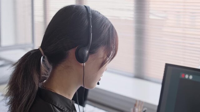 Chest up shot of female Asian operator in headset consulting customer while sitting at table in front of computer in modern call center