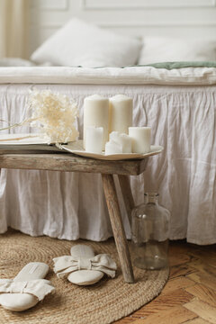 Close-up of assorted block candles and dried flowers on a wooden bench at the end of a bed with a pair of slippers and glass vase