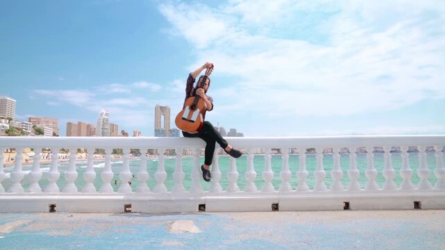 Flamenco artist picks up Spanish guitar to play with city and ocean background. Slow camera approach.