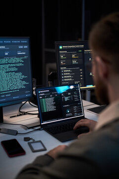 Vertical shot of young adult man monitoring cybersecurity dashboards across laptop and desktop screens, analyzing network data and threat alerts. Ideal for IT security, data protection, cyber defense