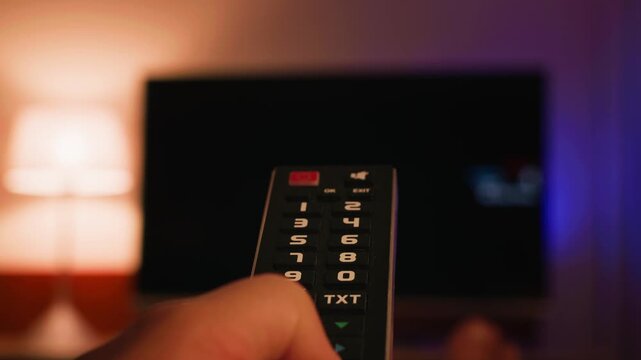 A close-up of a hand holding a TV remote control and changing channels. The TV screen is blurred in the background, creating a modern concept of home entertainment and relaxation. High quality 4k