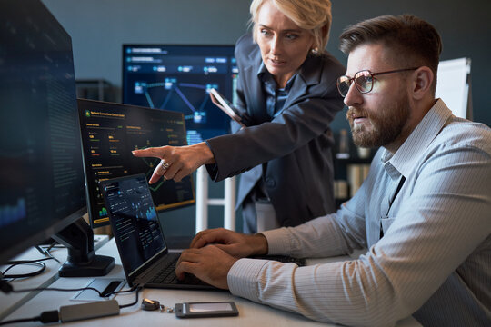 Caucasian businesswoman pointing at cybersecurity dashboard while Caucasian businessman analyzing data on laptop and monitors. Ideal for IT teamwork, network security, risk management