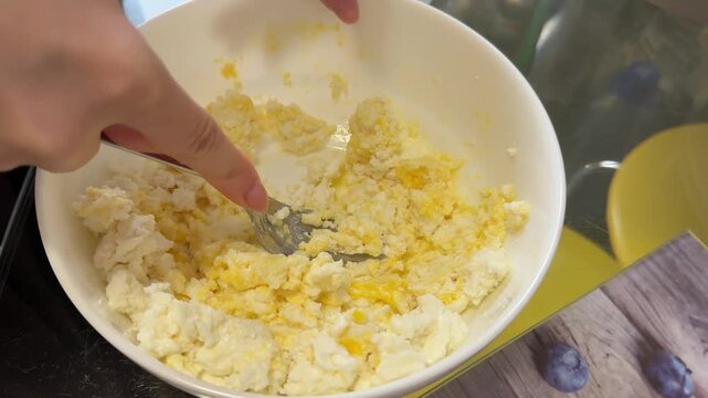Female hands adding fresh egg into cottage cheese mixture in white bowl while preparing ingredients for syrniki cheesecake or baked curd dessert.